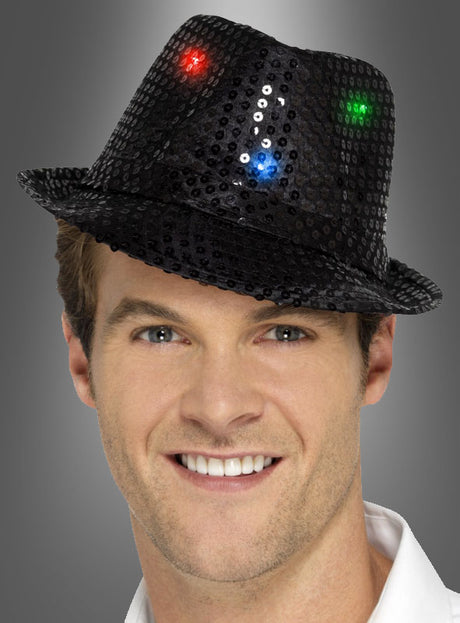 Eine lächelnde Person, die einen schwarzen Trilby-Hut mit LED trägt, der mit roten, blauen und grünen Lichtern verziert ist, vor einem grauen Hintergrund.