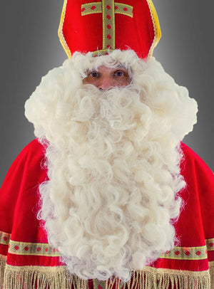 Eine Person, die als Nikolaus in einem rot-goldenen Gewand und einer Mütze verkleidet ist und den „Deluxe Bischof Nikolaus Bart und Perücke“ trägt, steht vor einem schlichten grauen Hintergrund.