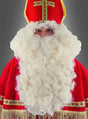 Eine Person, die als Nikolaus in einem rot-goldenen Gewand und einer Mütze verkleidet ist und den „Deluxe Bischof Nikolaus Bart und Perücke“ trägt, steht vor einem schlichten grauen Hintergrund.