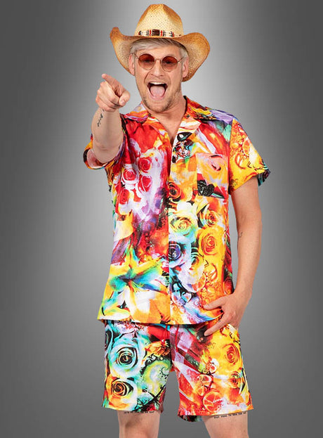Ein Mann, der ein buntes Hawaii-Hemd und eine bunte Hawaii-Hose trägt sowie einen Strohhut und eine Sonnenbrille, lächelt und zeigt vor einem grauen Hintergrund in die Kamera.