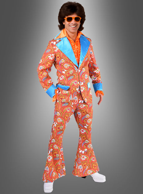 Eine Person, die den „Anzug Schlagerparty Martin“ trägt – einen farbenfrohen Paisley-Anzug im Stil der 1970er Jahre mit Schlaghosen, leuchtend blauen Revers, einem orangefarbenen Hemd, passender Sonnenbrille und weißen Plateauschuhen – ein kultiger 70er-Jahre-Anzug, ideal als Hippie-Anzug für Herren oder Schlagerparty-Kostüm – posiert lächelnd vor einem grauen Hintergrund.
