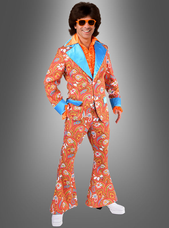 Eine Person, die den „Anzug Schlagerparty Martin“ trägt – einen farbenfrohen Paisley-Anzug im Stil der 1970er Jahre mit Schlaghosen, leuchtend blauen Revers, einem orangefarbenen Hemd, passender Sonnenbrille und weißen Plateauschuhen – ein kultiger 70er-Jahre-Anzug, ideal als Hippie-Anzug für Herren oder Schlagerparty-Kostüm – posiert lächelnd vor einem grauen Hintergrund.