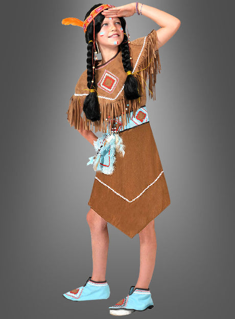 Ein Kind präsentiert das „Kleine Squaw“-Indianerkostüm, bestehend aus einem braunen Kleid im Wildleder-Look mit Fransen und Perlenstickerei, einem mit einer Feder verzierten Stirnband, geflochtenen Haaren und verzierten Schuhen, vor einem grauen Hintergrund.
