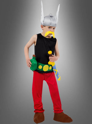 Ein Kind, das das „Deluxe Asterix Kinderkostüm Original“ trägt – komplett mit einem Wikingerhelm im Cartoon-Stil, einem gelben Schnurrbart, einem schwarzen Oberteil, einer roten Hose, braunen Schuhen und verspielten Spielzeug-Accessoires –, steht selbstbewusst vor einem grauen Hintergrund.