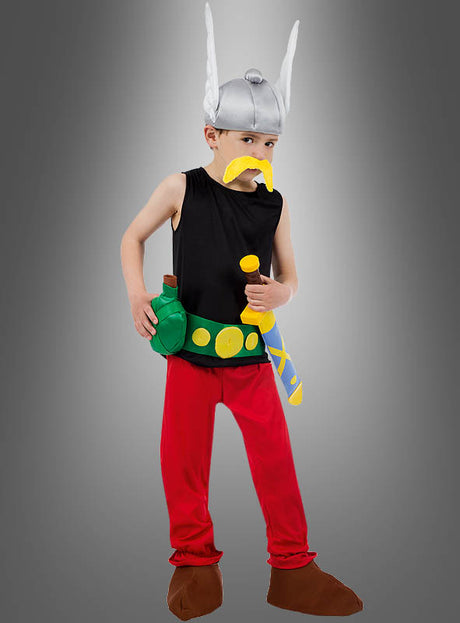 Ein Kind, das das „Deluxe Asterix Kinderkostüm Original“ trägt – komplett mit einem Wikingerhelm im Cartoon-Stil, einem gelben Schnurrbart, einem schwarzen Oberteil, einer roten Hose, braunen Schuhen und verspielten Spielzeug-Accessoires –, steht selbstbewusst vor einem grauen Hintergrund.