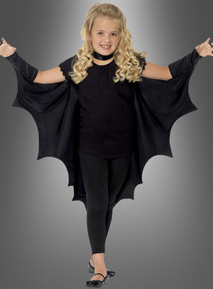 Vor einem grauen Hintergrund steht ein kleines Mädchen, das den „Fledermaus Vampir Umhang für Kinder“ trägt, die Flügel ausgebreitet, dazu schwarze Leggings und Schuhe – eine bezaubernde Wahl für Kinderkostüme oder Halloween-Spaß.