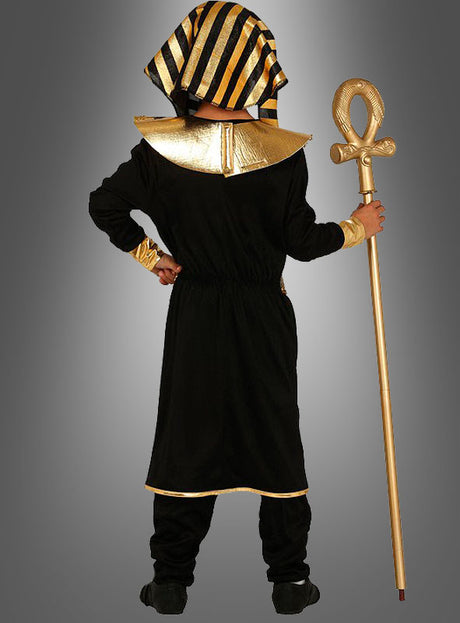 Ein Kind in einem schwarz-goldenen Pharao-Kostüm für Kinder steht mit dem Rücken zur Kamera und hält einen goldenen Stab mit einer Ankh-förmigen Spitze vor einem grauen Hintergrund mit Farbverlauf – ein Bild, das die majestätische Ausstrahlung dieses ägyptischen Kostüms perfekt einfängt.