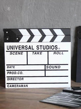 Auf einer Holzunterlage liegt eine schwarz-weiße Filmklappe als Hollywood-Partyzubehör mit Feldern für Szene, Take, Rolle, Datum, Ton, Produktion, Regisseur und Kameramann – damit ist sie das perfekte Kostümzubehör für jede Hollywood-Party.