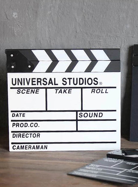 Auf einer Holzunterlage liegt eine schwarz-weiße Filmklappe als Hollywood-Partyzubehör mit Feldern für Szene, Take, Rolle, Datum, Ton, Produktion, Regisseur und Kameramann – damit ist sie das perfekte Kostümzubehör für jede Hollywood-Party.