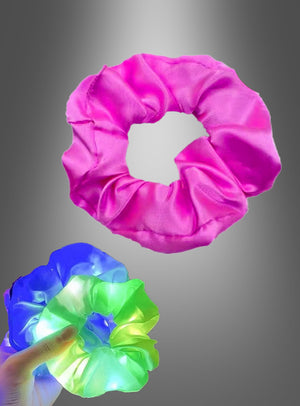 Eine Hand hält ein leuchtendes Scrunchie-Haargummi mit LED, dessen blaue, grüne und gelbe Lichter neben einem schlichten rosa Scrunchie vor einem Hintergrund mit Farbverlauf hell leuchten.