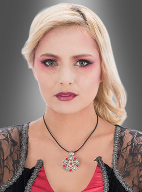 Eine Frau mit blonden Haaren, die ein auffälliges rosa Make-up trägt, eine auffällige „Pentagramm“-Halskette aus Silber mit roten Steinen, ein rotes Oberteil und einen schwarzen Spitzenüberwurf, posiert vor einem grauen Hintergrund mit Farbverlauf.