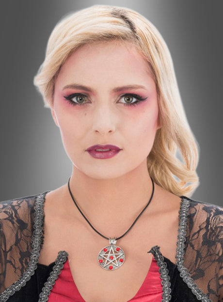 Eine Frau mit blonden Haaren, die ein auffälliges rosa Make-up trägt, eine auffällige „Pentagramm“-Halskette aus Silber mit roten Steinen, ein rotes Oberteil und einen schwarzen Spitzenüberwurf, posiert vor einem grauen Hintergrund mit Farbverlauf.