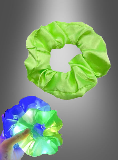 Eine Hand hält ein leuchtend blau-grünes Scrunchie-Haargummi mit LED, während ein weiteres Scrunchie-Haargummi mit LED in schlichtem Grün darüber vor einem grau verlaufenden Hintergrund schwebt.