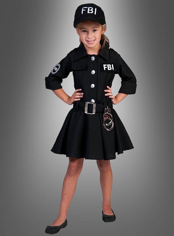 Vor einem grauen Hintergrund steht ein junges Mädchen lächelnd, gekleidet in eine FBI-Uniform für Mädchen, komplett mit Rock, Mütze, Dienstmarke und Handschellen.