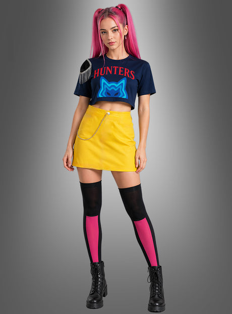 Eine Frau mit rosa Haaren in Zöpfen trägt das „K-Pop Hunters Mari“-Outfit für Damen – bestehend aus einem marineblauen Crop-Top, einem gelben Rock mit Kette, schwarzen Stiefeln sowie schwarzen und rosa Overknee-Socken – und posiert vor einem grauen Hintergrund, was einen auffälligen, von K-Pop-Sängerinnen inspirierten Look ergibt.