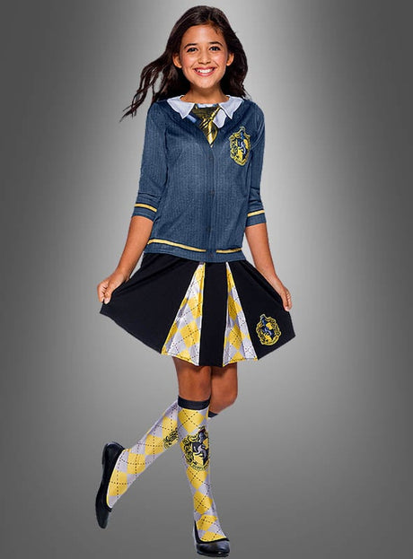 Ein lächelndes Mädchen trägt das „Kinder Hufflepuff“-Shirt aus der Harry-Potter-Reihe sowie eine blaue Jacke, einen gelb-schwarz karierten Rock, eine dazu passende Krawatte und Socken sowie schwarze Schuhe und steht vor einem grauen Hintergrund – ihr Outfit erinnert an eine klassische Hogwarts-Schuluniform und eignet sich ideal als Harry-Potter-Kostüm für Mädchen.