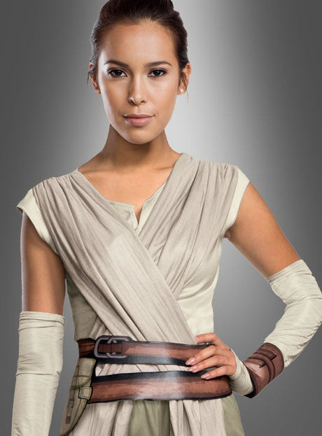 Eine Person, die das „STAR WARS: Episode 7 – Rey“-Kostüm Deluxe trägt – ein beiges Wickelkleid mit braunen Gürteln und Armwickeln –, steht vor einem grauen Hintergrund mit Farbverlauf.