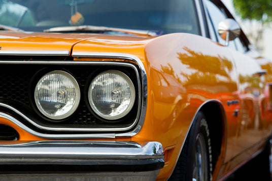 Nahaufnahme der vorderen linken Seite eines orangefarbenen Oldtimers, auf der die Scheinwerfer, der Kühlergrill sowie ein Teil der Motorhaube und des Seitenspiegels zu sehen sind.