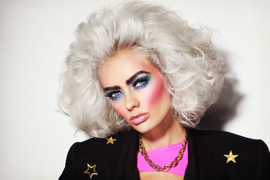 Eine Person mit voluminösem weißem Haar, auffälligem, farbenfrohem Make-up und einem schwarzen Blazer mit Sternenaufnähern, die eine goldene Halskette und ein rosa Oberteil trägt und vor einem schlichten Hintergrund posiert.