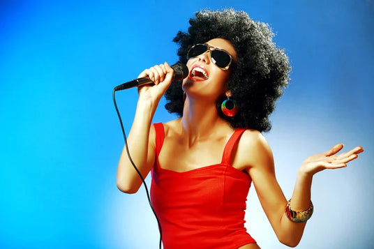 Eine Frau in einem roten Kleid mit einer Afro-Perücke und einer Sonnenbrille, die vor einem blauen Hintergrund in ein Mikrofon singt.