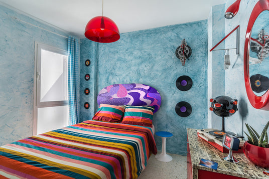 Ein farbenfrohes Schlafzimmer mit einer gestreiften Tagesdecke, einem gemusterten Kopfteil, hellblauen strukturierten Wänden, Schallplatten an der Wand, roten Akzenten und einem Fenster mit blauen Vorhängen.