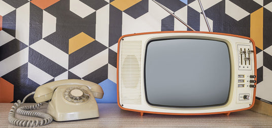 Auf einem Tisch vor einer Wand mit geometrischem Muster stehen ein altes Wählscheibentelefon und ein altmodischer Fernseher.