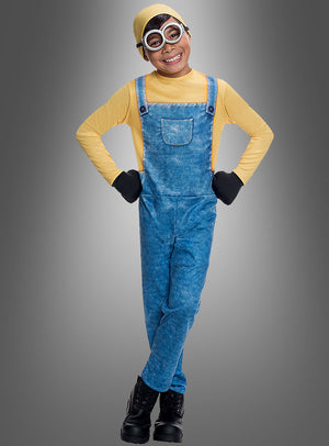 Ein Kind, das das Minion-Kostüm „Bob für Kinder“ trägt – mit einem gelben Hemd, einer blauen Jeanslatzhose, schwarzen Handschuhen, einer Schutzbrille, einem gelben Hut und schwarzen Stiefeln –, steht lächelnd mit den Händen in die Hüften gestemmt vor einem schlichten grauen Hintergrund und verkörpert damit perfekt den verspielten Charakter dieses Kostüms.