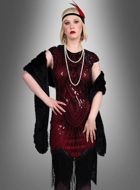 Eine Frau, die das „Hochwertiges Gatsby Kleid Daisy“ trägt – ein rot-schwarzes Kleid im Stil der 1920er Jahre mit Fransen –, ergänzt durch eine lange Perlenkette, ein Haarreif mit Federn und einen schwarzen Schal, steht vor einem grauen Hintergrund mit Farbverlauf.