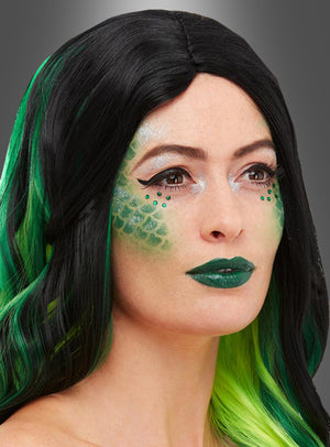 Eine Frau mit langen schwarzen und grünen Haaren, grünem Lippenstift und schuppigem grünem Make-up auf den Wangen – aufgebracht mit der Schuppen-Schablone aus dem Reptil-Make-up-Set mit Schuppen-Schablone – blickt vor einem grauen Hintergrund nach vorne.