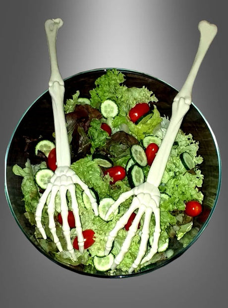 Eine Glasschüssel mit Salat aus Blattsalat, Kirschtomaten und Gurkenscheiben wird mit zwei „Salatbesteck Skeletthände“-Skeletthänden und -armen garniert.
