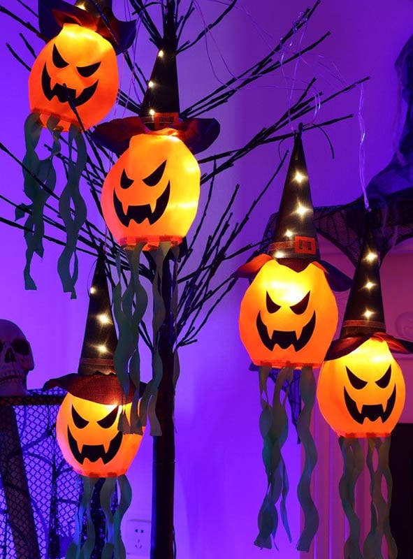 Fünf beleuchtete Kürbislaternen mit Hexenhüten hängen an den Ästen und sind mit grünen Papierschlangen sowie der Halloween-Girlande 280 cm mit LED-Kürbis oder Gespenst geschmückt, wodurch vor einem violett beleuchteten Hintergrund ein festlicher Halloween-Dekoeffekt entsteht.