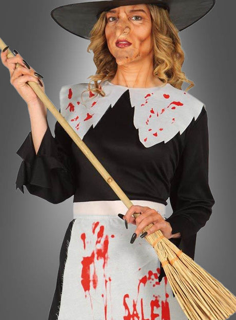 Eine Person in einem Hexenkostüm mit schwarzem Hut, die einen Hexenbesen für Zauberer und Hexen als klassisches Halloween-Accessoire in der Hand hält und ein schwarzes Kleid sowie eine weiße Schürze trägt, die mit roten Flecken und dem Wort „Salem“ verziert ist.