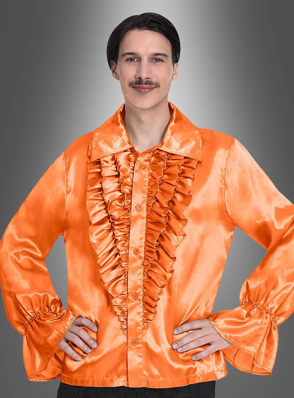 Eine Person, die das orangefarbene Retro-70er-Rüschenhemd „Schlagerstar“ mit Rüschen an der Vorderseite und ausgestellten Ärmeln trägt, steht vor einem grauen Hintergrund mit Farbverlauf. Die glänzende Satin-Oberfläche des Hemdes unterstreicht dessen vintage-inspirierten „Schlagerstar“-Stil.