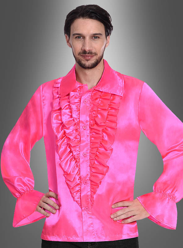 Ein Mann steht mit den Händen in den Hüften da und trägt das Retro-70er-Rüschenhemd „Schlagerstar“ in leuchtendem Pink, das vorne mit Rüschen und ausgestelltem Ärmelbund verziert ist und vor einem grauen Farbverlauf den kühnen Stil der 70er Jahre verkörpert.