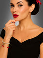 Eine Frau mit rotem Lippenstift, geschwungenem Eyeliner und einer roten Schleife im Haar, die ein schwarzes schulterfreies Kleid und das goldene Strass-Armband mit Kirschen trägt und mit der Hand am Kinn posiert.