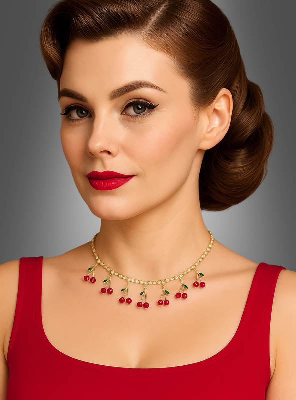 Eine Frau mit braunen Haaren, die zu einer Vintage-Hochsteckfrisur gestylt sind, trägt ein rotes Kleid und die „Strass Kette mit Kirschen 50er“ – eine goldene Halskette, die mit kleinen Kirschanhängern verziert ist –, während sie vor einem grauen Hintergrund posiert.