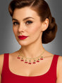 Eine Frau mit braunen Haaren, die zu einer Vintage-Hochsteckfrisur gestylt sind, trägt ein rotes Kleid und die „Strass Kette mit Kirschen 50er“ – eine goldene Halskette, die mit kleinen Kirschanhängern verziert ist –, während sie vor einem grauen Hintergrund posiert.