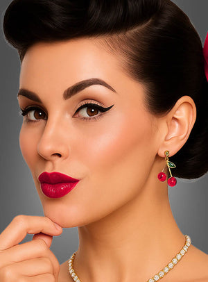 Eine Frau mit einer Retro-Frisur, rotem Lippenstift, einem geschwungenen Eyeliner und einer Perlenkette posiert vor einem grauen Hintergrund und trägt die Ohrringe mit Kirschen-Anhänger.