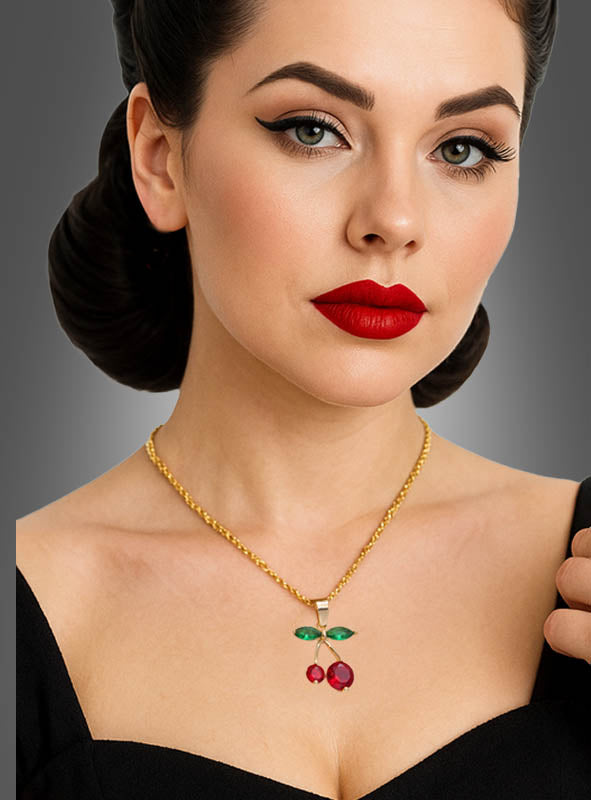 Eine Frau mit Retro-Make-up und -Frisur trägt die „Halskette mit Kirschen-Anhänger“ – eine goldene Kette mit einem Kirschanhänger, der mit grünen und roten Edelsteinen verziert ist – und posiert vor einem schlichten grauen Hintergrund.