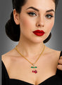 Eine Frau mit Retro-Make-up und -Frisur trägt die „Halskette mit Kirschen-Anhänger“ – eine goldene Kette mit einem Kirschanhänger, der mit grünen und roten Edelsteinen verziert ist – und posiert vor einem schlichten grauen Hintergrund.