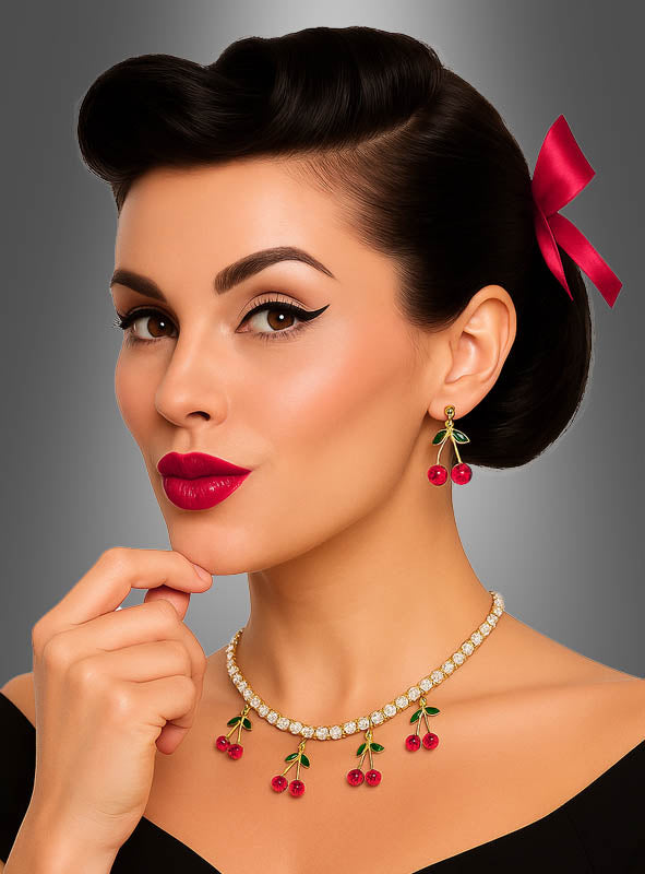 Eine Frau mit einer Retro-Frisur und einer roten Schleife präsentiert die Ohrringe mit Kirschen-Anhänger, kombiniert mit einer passenden Halskette, einem schwarzen Oberteil und einem geflügelten Eyeliner, und posiert vor einem grauen Hintergrund mit Farbverlauf.