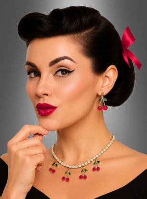 Eine Frau mit einer Retro-Frisur und einer roten Schleife präsentiert die Ohrringe mit Kirschen-Anhänger, kombiniert mit einer passenden Halskette, einem schwarzen Oberteil und einem geflügelten Eyeliner, und posiert vor einem grauen Hintergrund mit Farbverlauf.