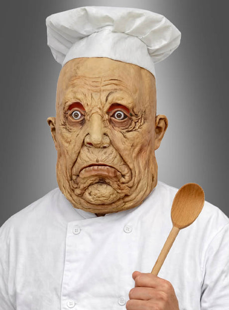 Eine Person in Kochuniform trägt die „Böse Zombie-Koch-Vollmaske“ – eine zerknitterte, übertrieben gestaltete Maske mit finsterem Blick – und hält einen Holzlöffel vor einem grauen Hintergrund in der Hand.