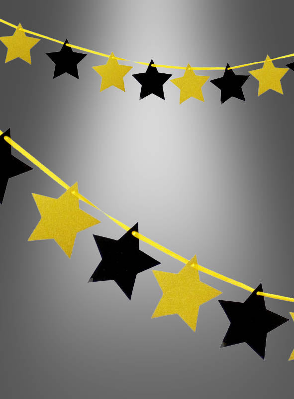Vor einem grau verlaufenden Hintergrund hängen zwei „Sterngirlanden“ in Schwarz-Gold mit Glitzer (300 cm).