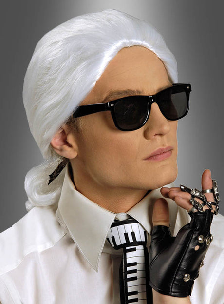 Eine Person, die die Perücke „Karl“ von Modeschöpfer trägt, eine schwarze Sonnenbrille, ein weißes Hemd mit Kragen, eine Krawatte mit Klaviertastenmuster und einen schwarzen Handschuh mit Metallfingern, posiert vor einem grauen Hintergrund mit Farbverlauf.