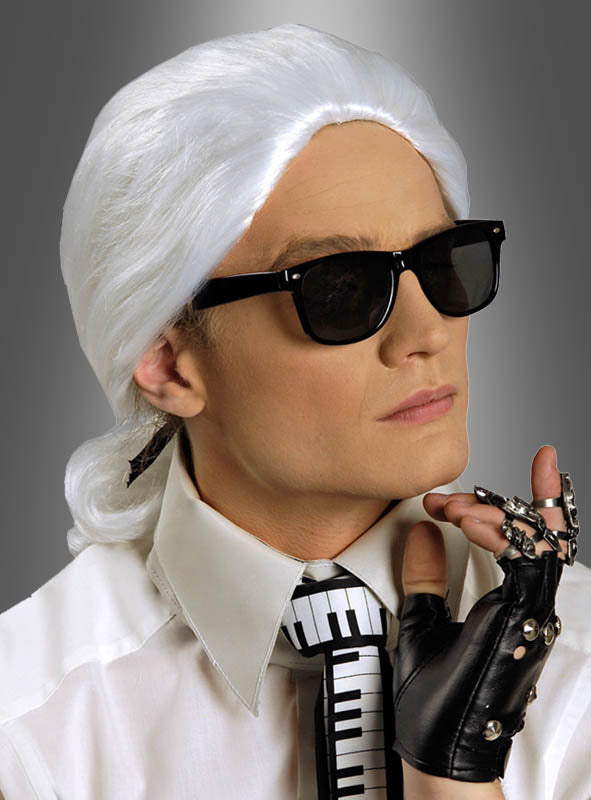Eine Person, die die Perücke „Karl“ von Modeschöpfer trägt, eine schwarze Sonnenbrille, ein weißes Hemd mit Kragen, eine Krawatte mit Klaviertastenmuster und einen schwarzen Handschuh mit Metallfingern, posiert vor einem grauen Hintergrund mit Farbverlauf.