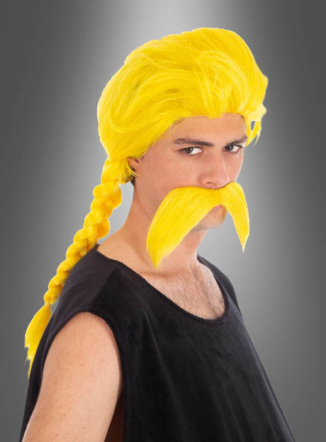 Ein Mann mit einem langen, gelben, geflochtenen Verleihnix-Bart und einer Perücke, gekleidet in ein ärmelloses schwarzes Oberteil, posiert vor einem grauen Hintergrund mit Farbverlauf in einem auffälligen Asterix-Kostüm.