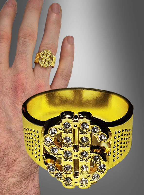 Eine Nahaufnahme einer Hand, an der der „Dollar Ring Herren“ steckt, ein Goldring mit einem Dollarzeichen-Motiv, das mit klaren Steinen verziert ist.