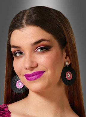 Eine Frau mit glattem braunem Haar, leuchtend rosa Lippenstift und auffälligem Augen-Make-up trägt die „50er Ohrringe Schallplatte schwarz-pink Clips“ – schwarze Clip-Ohrringe in Form einer Schallplatte – vor einem grauen Hintergrund mit Farbverlauf.