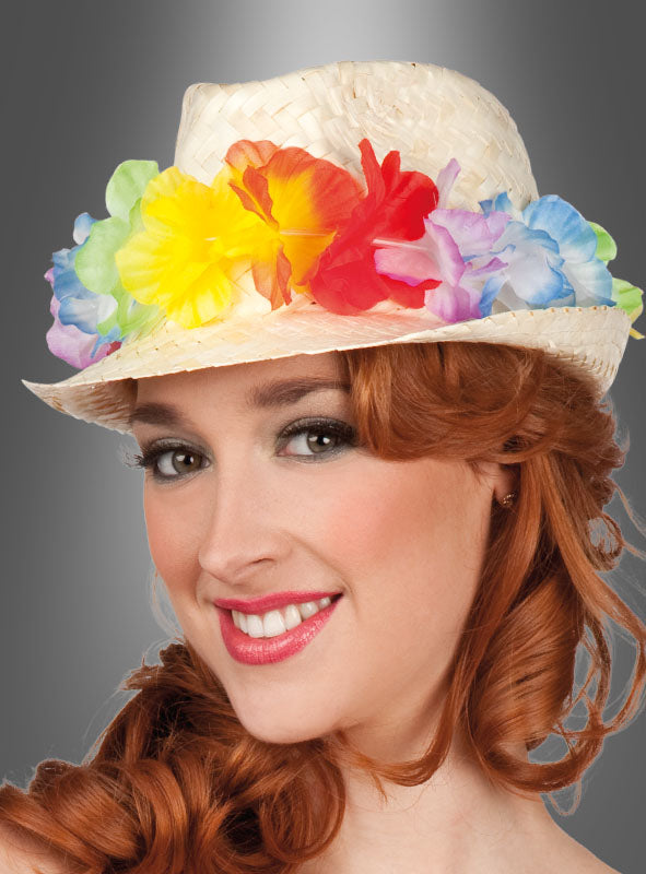 Vor einem grauen Hintergrund lächelt eine rothaarige Frau, die einen „Strohhut mit Blumenkette“ trägt – einen Strohhut, der mit bunten Kunstblumen verziert ist.